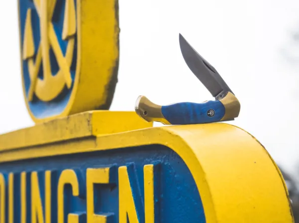 Nóż Böker Scout Solingen Während der Feierlichkeiten anlässlich des 150-jährigen Jubiläums der Böker Manufaktur fragte uns der Oberbürgermeister der Stadt Solingen, ob wir ein hochwertiges Taschenmesser in den Farben der Stadt entwickeln könnten. Er würde sich sehr freuen, ein solches Messer zu offiziellen Anlässen an Gäste überreichen zu können. Diese Anfrage ehrte uns sehr und schnell fiel die Wahl für das Grundmodell auf das überaus beliebte und vielseitige Böker Scout. Seit über dreißig Jahren überzeugt das Scout mit seiner eleganten und zeitlosen Formgebung sowie seiner stabilen und ergonomischen Bauweise mit gesintertem Griffkörper Messerfreunde auf der ganzen Welt. Die Farben der Stadt Solingen sind ein kräftiges Stahlblau und Goldgelb. Statt mit einem gesinterten Körper aus Neusilber wird das Scout Solingen erstmalig mit einem Körper aus massivem Messing ausgestattet, der präzise per CNC-Technik aus dem Vollen gefräst wird. In Anlehnung an den gewaltigen Kastanienbaum vor der ersten Böker Handwerkzeug-Fabrik, haben wir uns für Griffschalen aus blau stabilisiertem Holz der Rosskastanie entschieden. Um die jahrhundertealte Tradition der Klingenstadt Solingen zu betonen, wird die Klinge aus nicht rostfreiem C75 Kohlenstoffstahl gefertigt und mit einem authentischen Acid Wash Finish versehen. Die Auslieferung des komplett von Hand in der Böker Manufaktur Solingen gefertigten Messers erfolgt mit dem neu aufgelegten historischen Böker Baumzeichen-Emblem. Zudem möchten wir mit diesem besonderen Messer ein altes Solinger Ritual wiederaufleben lassen. Messer sind ein perfektes Präsent, über das sich fast jeder freut. Allerdings sagt der Volksmund in der Klingenstadt, dass ein verschenktes Messer das Band der Freundschaft zerschneidet. Um dieses Unglück zu verhindern, erhält der Beschenkte mit dem Messer eine Geldmünze, die er dem Schenker wieder zurückgibt. Dem Brauchtum folgend wurde das Messer so bezahlt und gilt nicht länger als unglücksverheißendes Geschenk. Damit das Böker Scout Solingen in Zukunft viele Freundschaften begründen und erhalten kann, wird jedes Messer daher mit einer praktischen Böker Münze ausgeliefert.