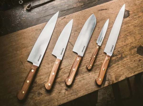 Nóż Santoku Böker Solingen Cottage-Craft Tak zwani Kotten kształtują historię miasta Solingen od XIV wieku. Masywne osełki napędzane były czystą wodą, a słynne na całym świecie ostrza szlifowano ręcznie. Seria noży szefa kuchni Böker Cottage-Craft imponująco kontynuuje dziedzictwo tego tradycyjnego rzemiosła i zachwyca starannie dobranym wyborem klasycznych materiałów. Na szczególną uwagę zasługuje zastosowanie rdzewnej stali węglowej C75, która po krótkim okresie użytkowania tworzy typową niebiesko-szarą patynę. Naturalne drewno śliwkowe użyte do wykonania muszli rękojeści jest twarde, gęste i drobnoporowate, ma jednolitą strukturę i uderzające spektrum kolorów. Zgodnie z wielowiekową tradycją produkcji noży z Solingen, łuski rękojeści są mocowane na ciągłym trzpieniu ostrza za pomocą nitów z litego mosiądzu.Nóż, który staje się coraz bardziej popularny w Europie, to Santoku. Pierwotnie był to japoński kształt ostrza noża użytkowego. W tłumaczeniu jego nazwa oznacza „Trzy cnoty” i nadaje się równie dobrze do mięsa, ryb i warzyw. Dzięki szerokiemu ostrzu Santoku jest szczególnie łatwy w obsłudze.CARBON STEEL C75 Całkowicie naturalną właściwością materiału jest to, że podczas używania noża powstaje patyna. Szaroniebieskie zmatowienie ostrza jest całkowicie normalne i nie jest w żaden sposób wadą. Wręcz przeciwnie, powstała w ten sposób patyna (warstwa tlenku) tworzy na ostrzu warstwę ochronną, która zwiększa jego odporność na warunki zewnętrzne. Ta patyna jest często widoczna już po pierwszym użyciu i nie można jej ponownie usunąć. Ekspert natychmiast rozpoznaje wynikającą z tego wysokiej jakości stal węglową. Przy odpowiedniej pielęgnacji stal węglowa ma taką samą żywotność jak nowoczesna stal nierdzewna. CZYSZCZENIE I PIELĘGNACJA Stal węglowa jest jednym z rdzewnych rodzajów stali i wymaga celowej, ale minimalnej konserwacji. W kuchni wystarczy opłukać nóż pod gorącą bieżącą wodą i osuszyć ściereczką. W przypadku dłuższego przechowywania ostrze należy konserwować odpowiednim olejem. Idealny do tego celu jest bezkwasowy, nieżywiczny i nielotny olej Boker Camellia (nr kat. 04BO175). Jest olejem czysto roślinnym i idealnie nadaje się do konserwacji rdzewnych noży kuchennych, w przeciwieństwie do innych olejów pielęgnacyjnych – nie tworzy emulsji z wodą. Z drugiej strony, używanie zmywarki niszczy drobne struktury stali węglowej, podobnie jak pozostawianie ostrza w zlewie na dłuższy czas bez czyszczenia. Rzadko stosowana w nożach kuchennych stal węglowa, ze względu na swoje właściwości można ją łatwo ostrzyć, dzięki czemu noże Boker Cottage Craft stają się lojalnymi i niezawodnymi towarzyszami na całe życie i kontynuują pisanie własnej historii.