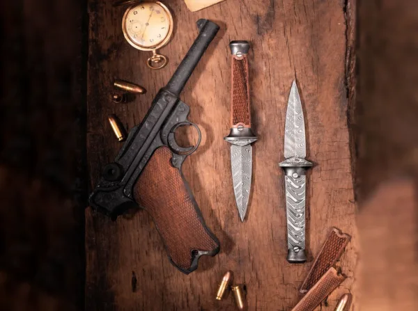 Nóż Böker Solingen P08-Damast The unique Boker P08-Damascus boot dagger connects the world of knives with historical military history in an inimitable way. The P08, also known as Parabellum Pistol or simply Luger, is still considered legendary today and convinces with highest precision and reliability. The knee-joint lock with short barrel return, developed by Georg Luger, made the pistol 08 incredibly compact and solid. Added to this was the calibre 9 mm Luger (9x19 NATO), also developed by Luger for the new pistol, which was one of the most powerful short firearm calibres at the time and is still the most widely used official calibre in the world today. It was used as a standard pistol by the German armies in both world wars, but also gained world fame beyond the country's borders in a very short time. As early as 1914, the weapon was used in the armies of Bulgaria, the Netherlands, Portugal and Turkey and was considered a much sought-after prey weapon until the end of the Second World War. The production of the compact pistol required 14 kilogrammes of steel and its design required such a high degree of precision that completely new demands were made on the production technology. Its unmistakable, almost iconic appearance secured it a firm place in popular culture like no other pistol. The Boker P08-Damascus not only incorporates various design elements of the unusually aesthetic official weapon, but is also equipped with a unique damascus blade made of the barrel steel of the precise pistol. The remarkable damascus is forged for us by Chad Nichols by hand in the Boomerang pattern. The handle scales of the likewise compactly built boot dagger are made of certified imported rosewood and feature the classic, simple fish-scale cut of the characteristic pistol grip. The same construction of the retaining screws is used to mount these handle scales as on the P08. And the cone of the exposed barrel is also found in the angle of the handle sides. The bolsters and end knob are made of steel and are finished with a dark stonewash finish. Supplied with a high quality black leather sheath with a sewn-in ulticlip and can be easily attached thanks to the locking spring clip. Thus the Boker P08-Damascus boot dagger can be pulled effortlessly while the sheath is reliably held in place by the Ulticlip. Handmade in the Boker Knife Manufactory in Solingen. With noble floating display, certificate of authenticity and individual serial number.