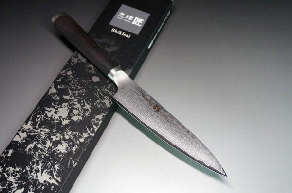 Shizu Hamono MIYAKO japoński nóż petty damast 13cm AUS-8 Nóż Uniwersalny Shizu Hamono Shikisai Miyako 130mm Nóż uniwersalny Shikisai Miyako SM-1060 o długości 130mm to wszechstronne narzędzie idealne do precyzyjnych zadań kuchennych. Doskonały do obierania, krojenia warzyw, owoców oraz delikatnych prac przy mięsie. Kompaktowy rozmiar zapewnia łatwość obsługi i codzienną funkcjonalność. Cechy Ostrza: Stal Damaskowana: 33 warstwy z rdzeniem ze stali AUS8. Skład Stopu: 0,8% węgla, 14,5% chromu. Wytrzymałość: AUS8 łączy twardość z elastycznością. Unikalne Cechy: Warstwy SUS410: Zwiększona wytrzymałość i równowaga ostrza. Młotkowanie Ostrza: Charakterystyczny wygląd. Rękojeść: Laminowane drewno, tradycyjny kształt "D" dla praworęcznych. Szczegóły Produktu: Producent: Shizu Hamono Co.,Ltd. (Japonia) Stal Ostrza: 33-warstwowa stal damasceńska, rdzeń AUS8 Twardość: 59 HRC Rękojeść: Laminowane drewno, stalowa nasadka Typ: Nóż uniwersalny (Petty/Utility Knife) Długość Ostrza: 130 mm Długość Całkowita: 235 mm Waga: 85 g Krawędź: Dwustronna (50/50) Kraj Produkcji: Japonia